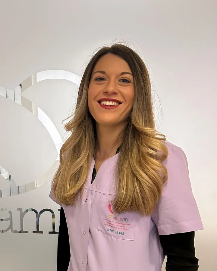 Eva, sonriendo, con el uniforme lila del centro junto al cristal grabado con el logotipo de AmuleTO