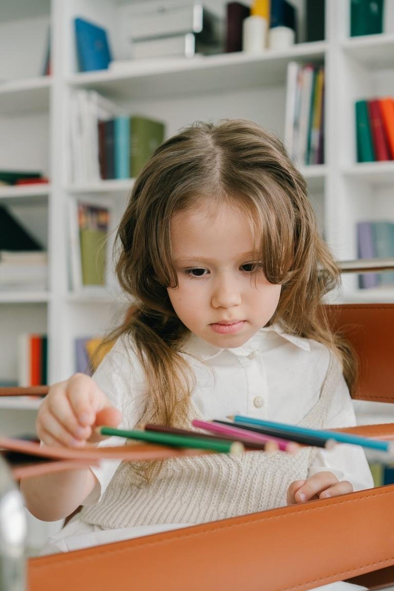 Niña sentada a la mesa con lápices de colores delante de una estantería